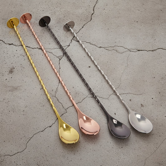 Four metallic spoons in gold, rose gold, silver, and bronze on a concrete surface.
